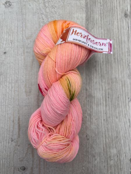 Handgefärbter Strang in Rosa- und Apricot-Tönen mit feinen Farbsprenkeln auf hellem Holzuntergrund; Herzfasern-Etikett sichtbar.“